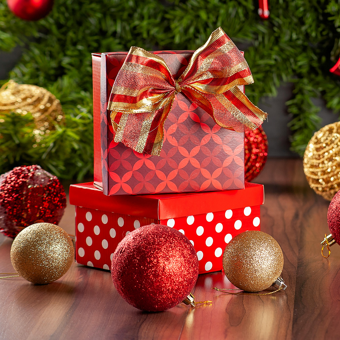 Two stacked red gift boxes with festive ribbon, surrounded by glittery red and gold Christmas ornaments and green garland on a wooden surface. Prefect gift for Christmas Holiday to someone you loved. Shop now.