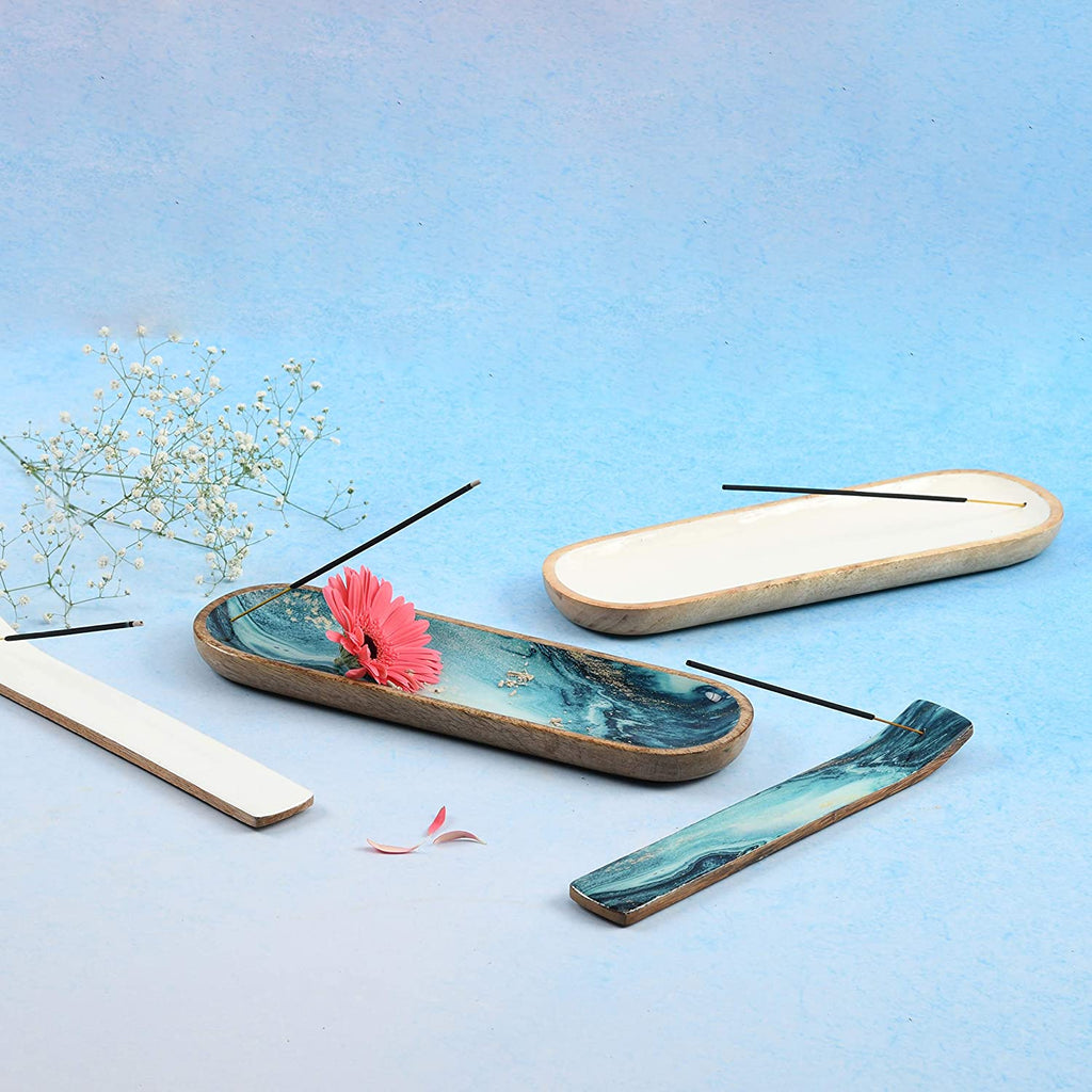 Set of incense holders—blue marble and white wood designs with burning sticks, pink flower, and scattered petals on light blue surface for calming decor