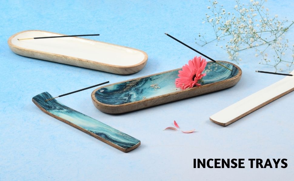 Collection of incense trays—blue marble, white ceramic, and wood designs with burning sticks, pink flower, and scattered petals on light blue surface