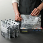 Person handling reusable aluminum foil seafood storage bags with labeled date and content fields, designed for freezing and refrigeration, shown with packaging box on kitchen counter.