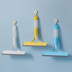 Three wall-mounted squeegees in white, yellow, and blue—hanging on adhesive hooks for organized storage and easy access to window cleaning tools